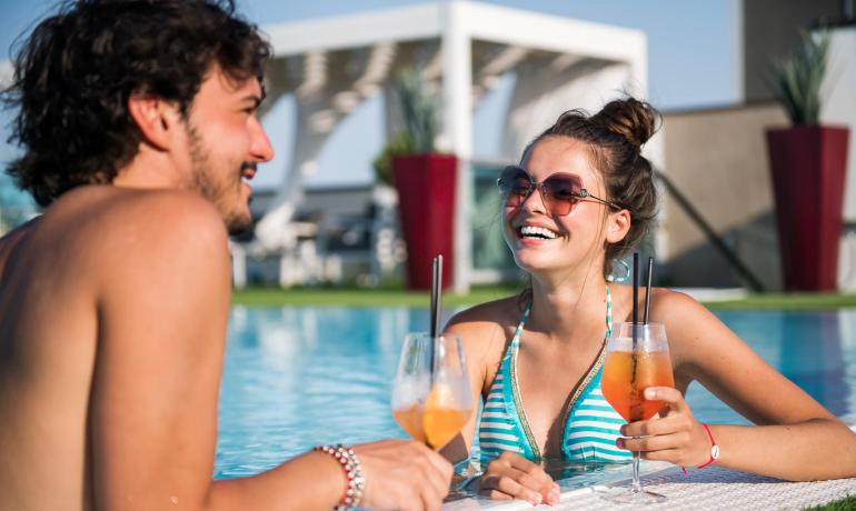 Due persone sorridenti in piscina con cocktail.